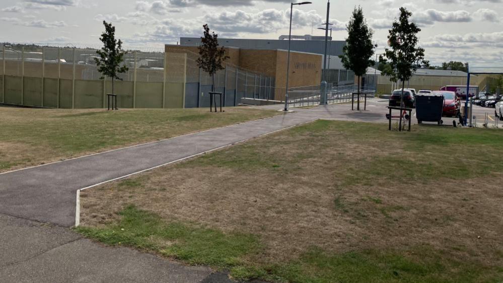 Path leading to prison building, which is surrounded by blue railings. A taller grey building is visible in the background. There are cars in a car park to the right. The path has grass on both sides.