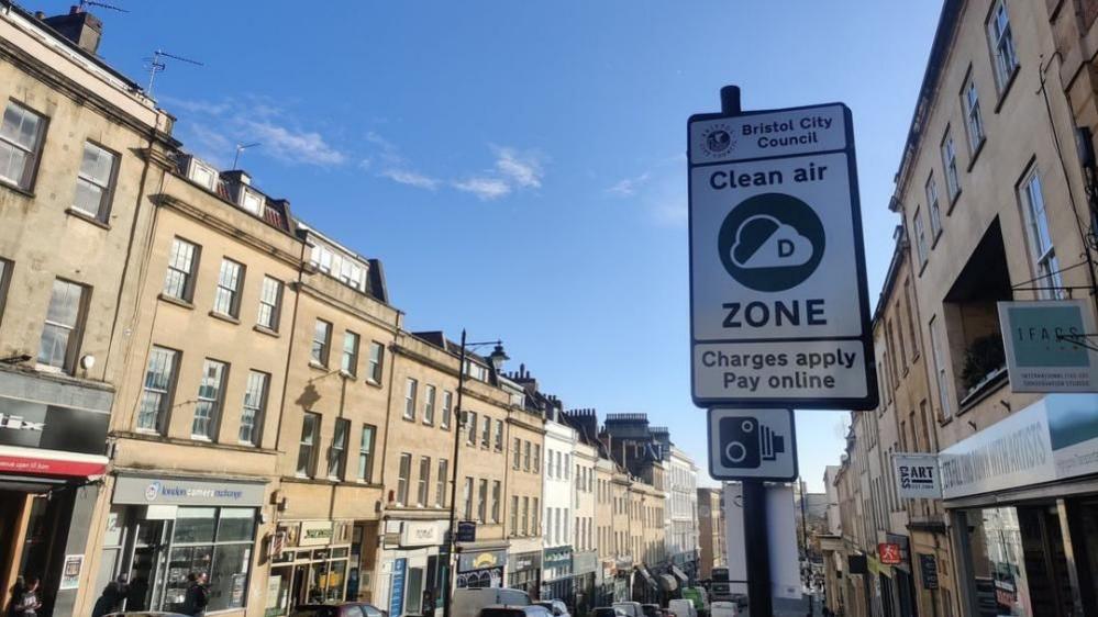 A row of houses and shops going down a steep hill. They are built of sand-coloured stone. A sign on the roadside reads 'Clean Air Zone, Charges Apply Online' and has a camera symbol on it.