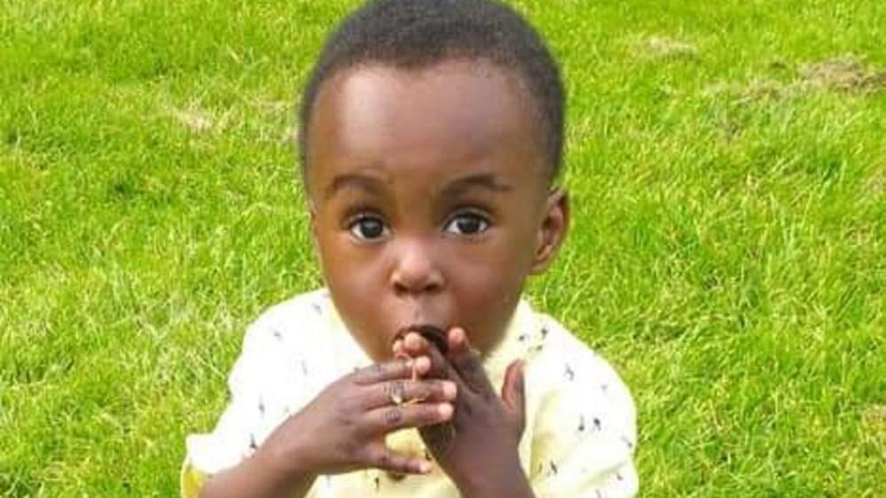 A little boy sits outside on some grass on a sunny day. He is holding his hand over his mouth.