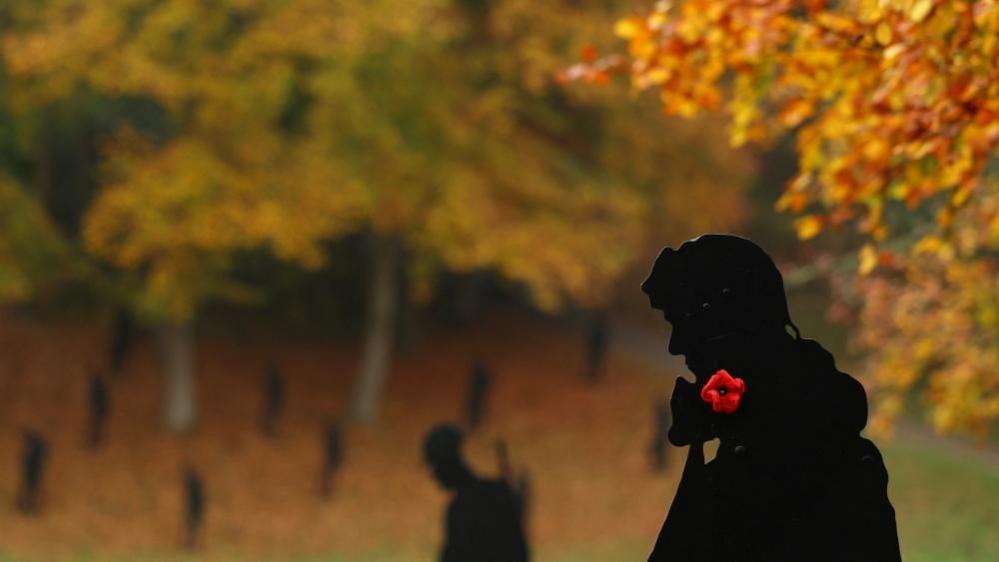 The silouettes of several servicemen in Stowe Gardens. One closest to the camera is a black figure with a red poppy on his neck. Autumn trees can be seen in the distance.