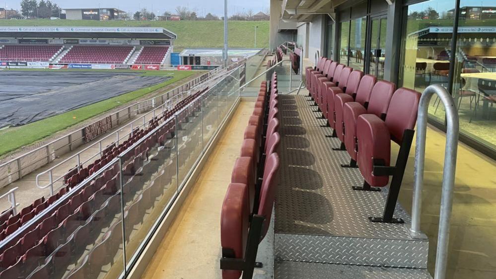 A view from the second level of a football stand. Two rows of claret seats are behind a glass balcony overlooking another row of seats and a covered football pitch. 