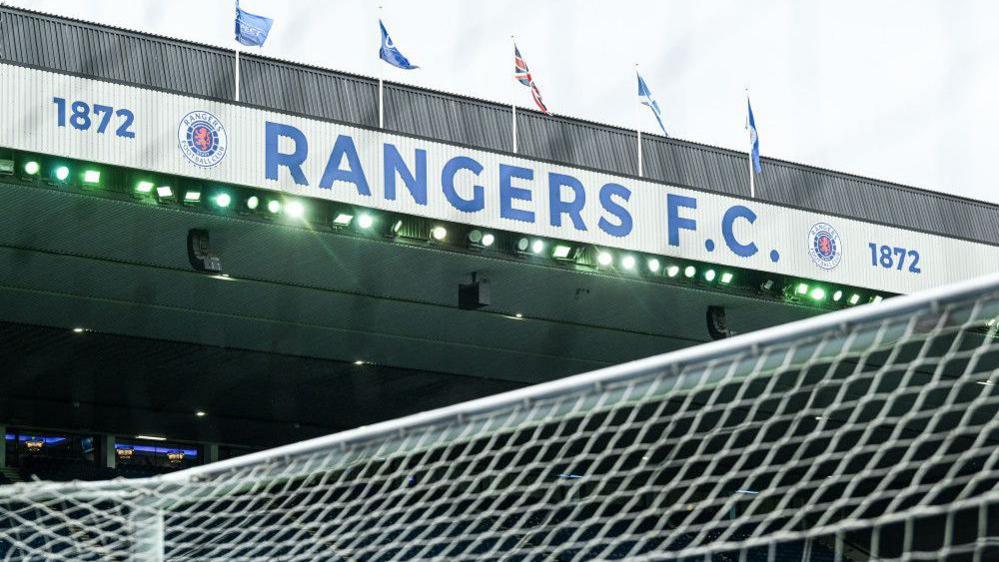 General view of Ibrox stadium under a grey sky showing the top of the goal and the main stand with flags fluttering in the wind above it