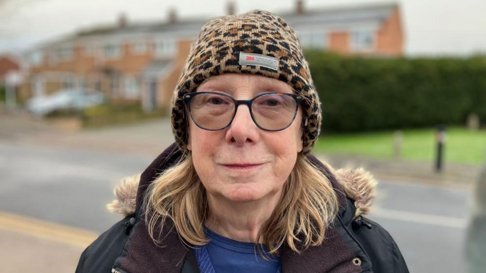 Ann Nevison standing in front of a row of houses and a green bus, which is slightly blurred. She is wearing a leopard print hat, with glasses and shoulder-length fair hair. She is wearing a black coat, with fur trim, a blue top and jumper.