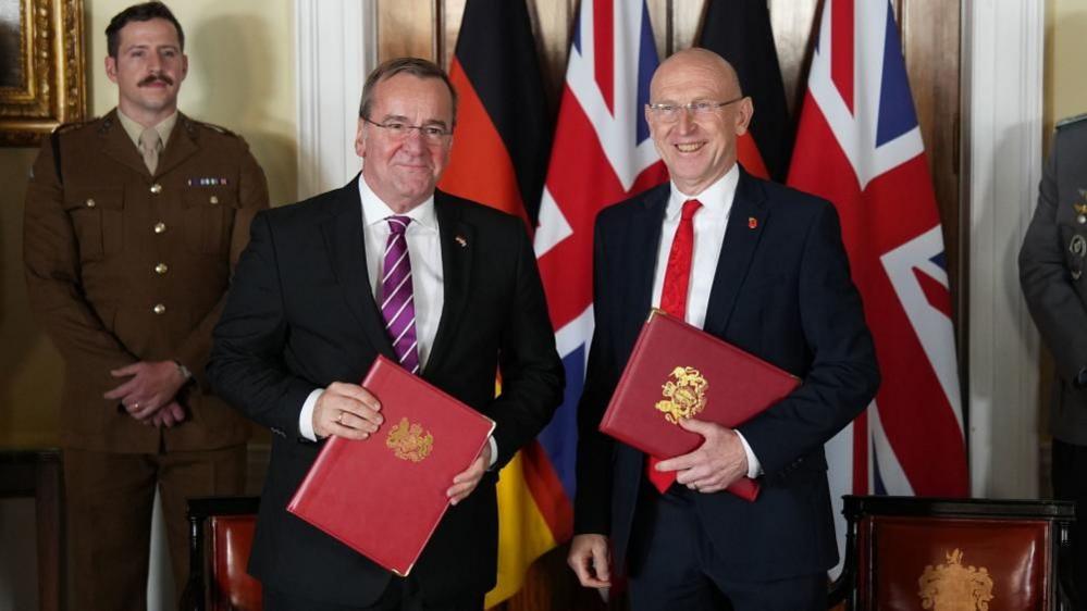 Healey and Pistorius smile, standing in front of the Union Jack and German flags, holding red binders 