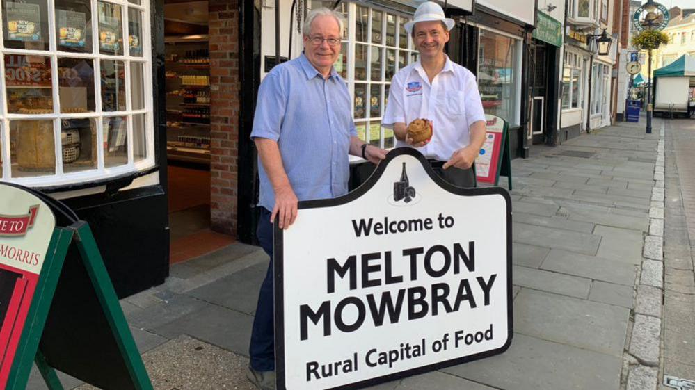 Matthew O'Callaghan (left) wearing a blue shirt next to a man from Dickinson & Morris pork pie bakery wearing a white shirt and hat. They are stood holding a white sign saying "Welcome to Melton Mowbray Rural Capital of Food.