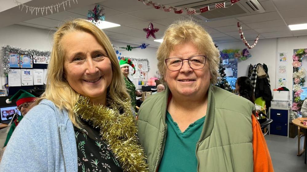 Diana Staines, right, is wearing a green top with orange sleeves, with a light green gilet over the top. She has light auburn hair, and is wearing glasses. Next to her, on the left of the image, is her successor, Alison Holmes, who is wearing a black patterned top beneath a light blue cardigan. She is wearing tinsel around her neck, and she has long auburn hair.