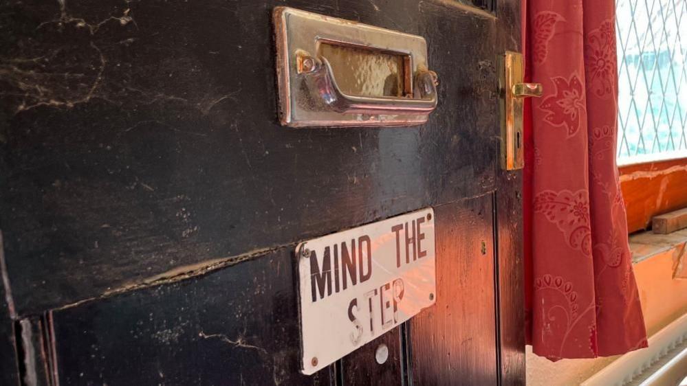 A "mind the step" sign affixed to the black-painted entrance door of the Decoy Tavern. A brass doorhandle and letterbox/door knocker combined are also mounted to the door. Next to it, red-coloured curtains border the window, and a radiator is visible beneath.