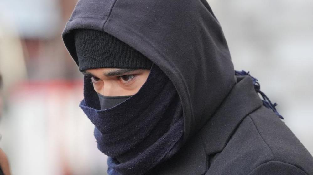Close up of Adeel Khan from the shoulders up. His face is largely obscured apart from his eyes and eyebrows by a black hat and face covering. He is also wearing a black scarf and black hoodie over his head.