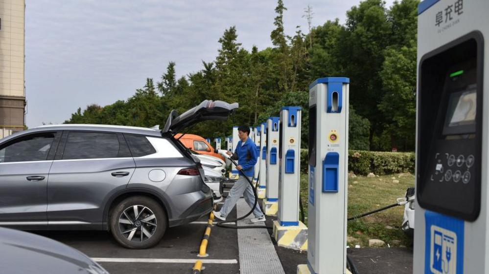 A man charges an electric car at an electric vehicle charging station on a street in Fuyang, China. Cars are parked in front of a row of blue and white charging stations.
