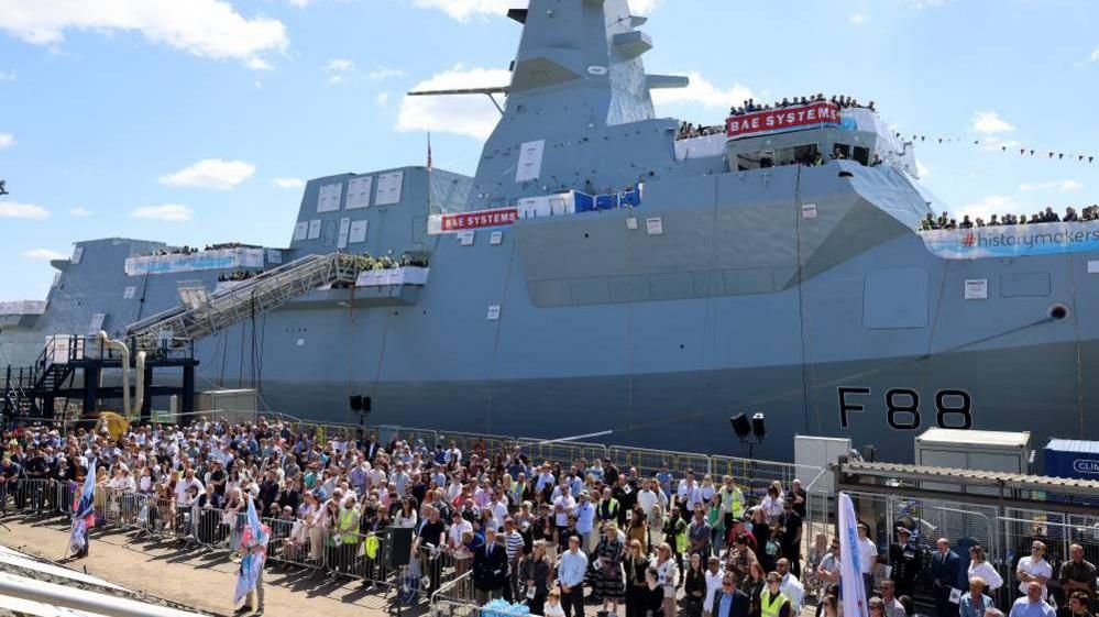 A large crowd gathered in front of a newly built warship. The ship is light grey and prominently displays "BAE Systems" branding. The crowd is largely dressed in business casual attire, suggesting an official launch or unveiling event. A banner reading "#historymakers" is visible on the ship.