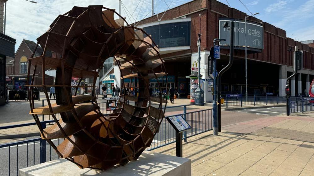 A vortex-shaped fan, made from Corten steel, mounted on a plinth on Regent Road and Alexandra Road, with Market Gates Shopping Centre in the backdrop.
