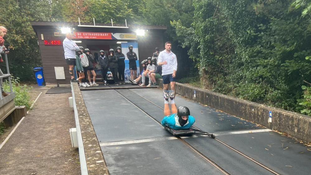 Someone heading down a skeleton track head first while a crowd of people watch in the background