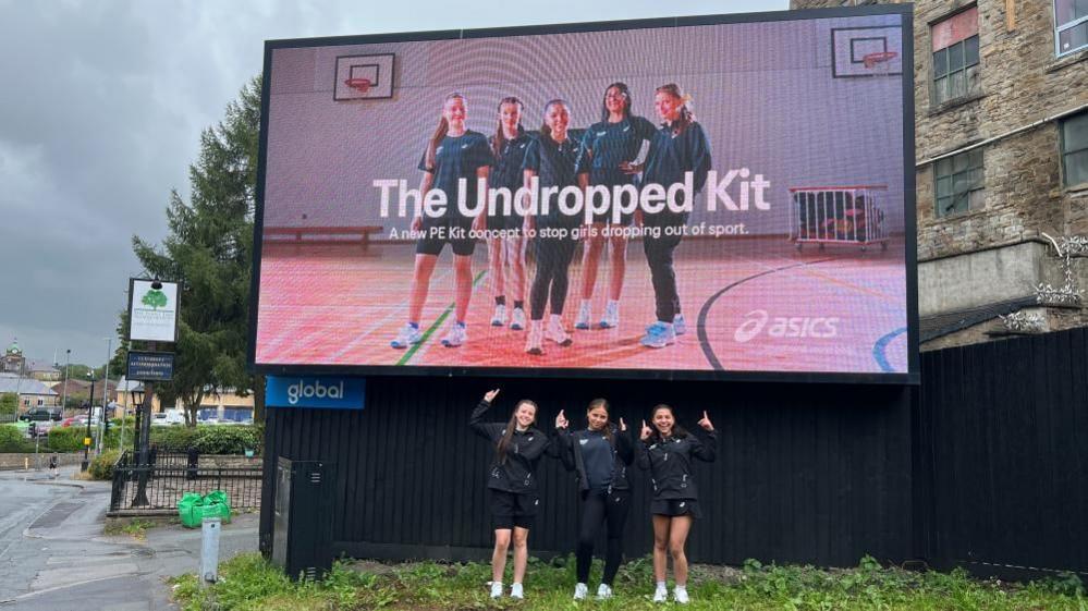 The three girls standing on a patch of grass, pointing their fingers upwards underneath an electronic billboard where the image for 'The Undropped Kit' is displayed, which they feature in along with two more of their classmates. It reads: The Undropped Kit, a new PE kit concept to stop girls dropping out of sport.