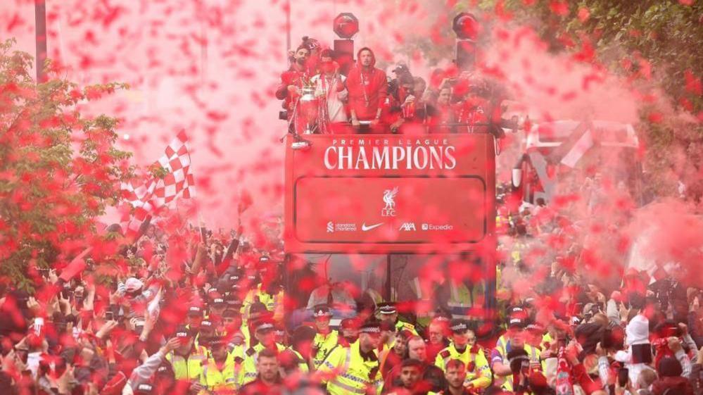 A red open-top bus carrying Liverpool football players drives through the city surrounded by crowds and red flares and smoke. There are several police officers at the front of the bus. 