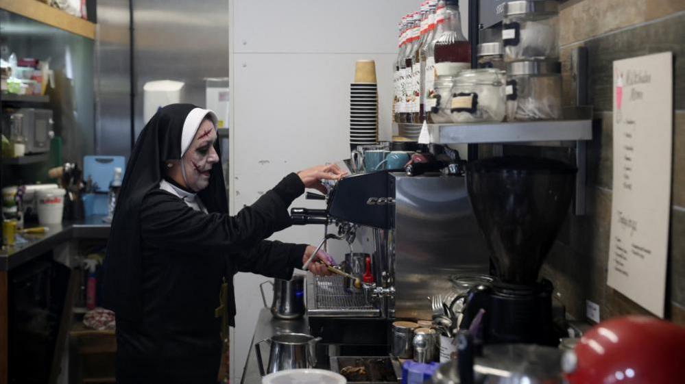 A person wearing a nun’s habit is operating a professional espresso machine in a small café setting. The counter is filled with coffee-making equipment, including a grinder, jars of ingredients, and stacked paper cups. Shelves above hold bottles of syrup and containers, and a menu is visible on the wall to the right.
