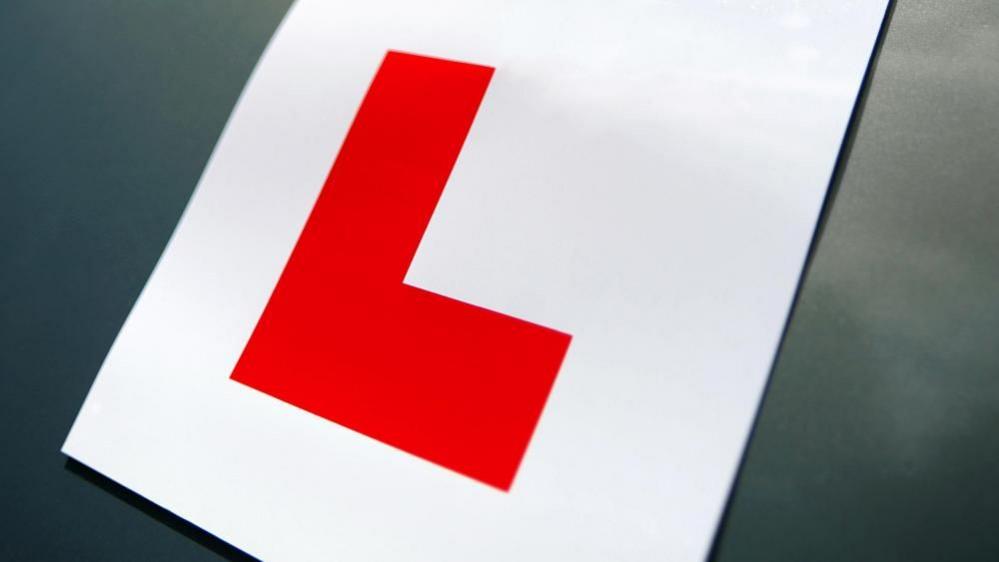 A close-up of an L plate. A large red capital L on a white background, fixed to a dark-coloured car.