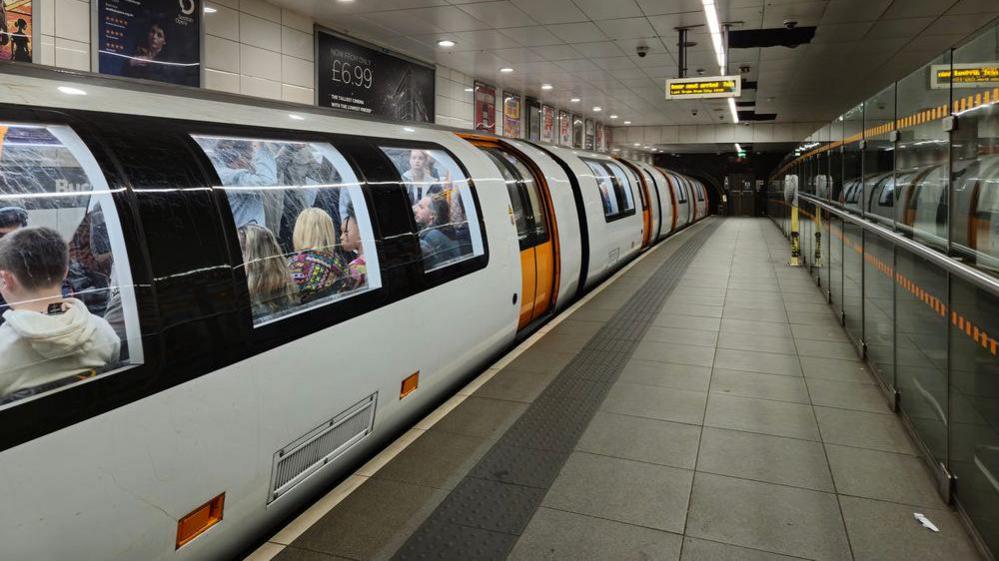 One of the new Glasgow subway trains is stationary on a platform. The train is white with orange doors and it is full of people. There is no one on the platform and the train doors are closed. Adverts can be seen behind the train.