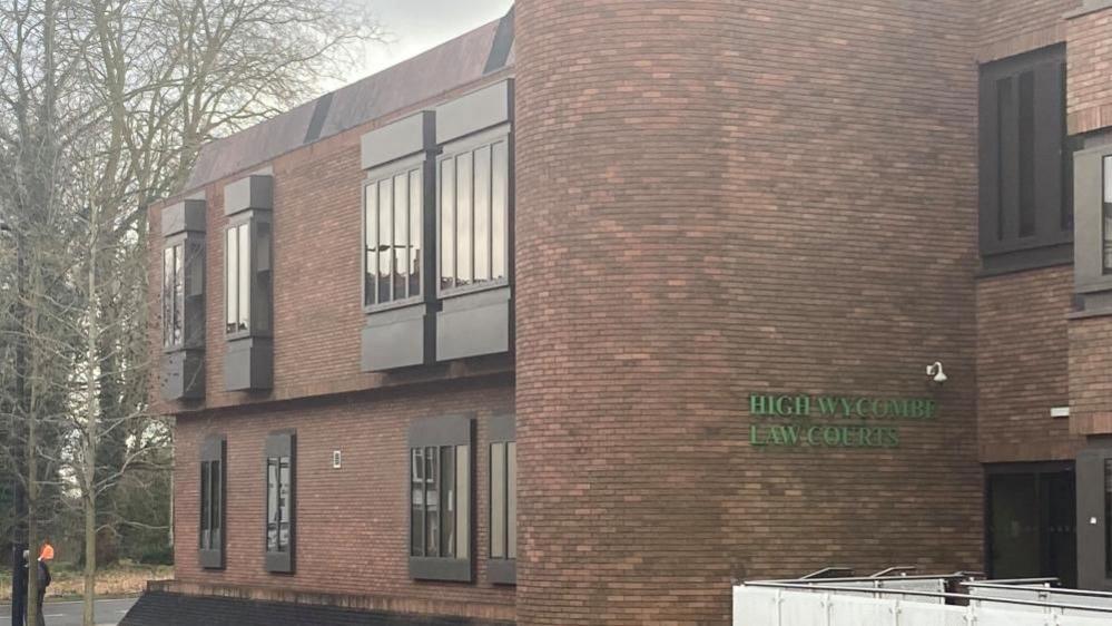 High Wycombe Law Courts: A red brick building with dark grey windows. The words "HIGH WYCOMBE LAW COURTS" are written in green capital letters on one wall. Trees and a road are visible on the left of the image. 