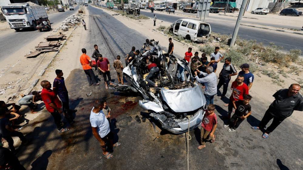 A large group of men surround a destroyed car