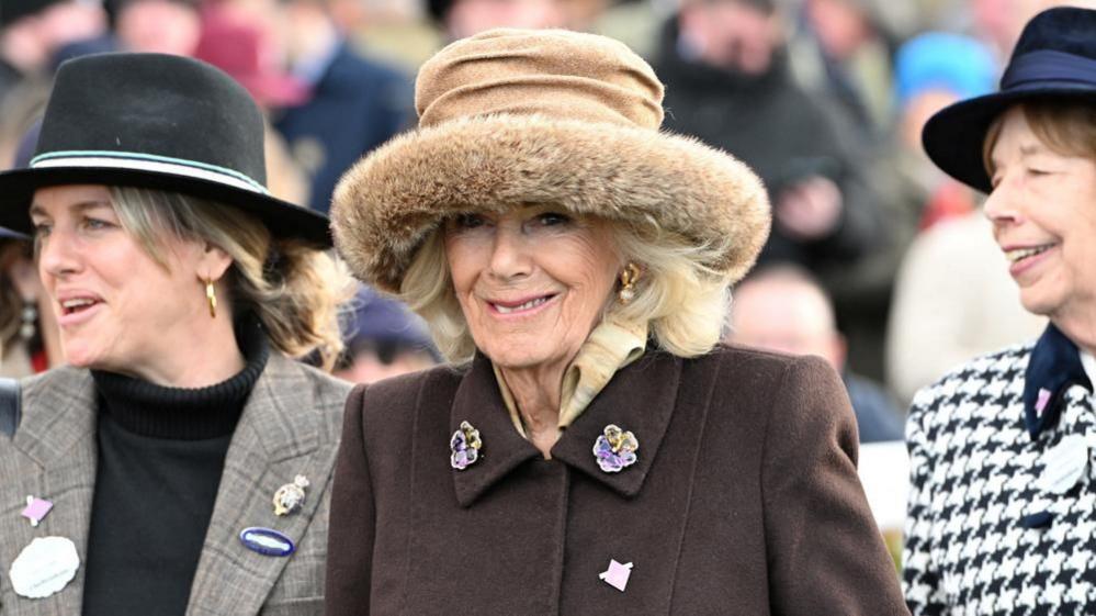 Queen Camilla, wearing a brown coat and brown hat, smiling towards the camera. She is in between two other women who are wearing hats and smiling. 