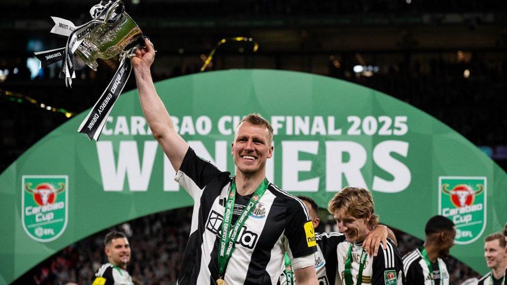Dan Burn after winning the Carabao Cup. He's wearing a black and white Newcastle United top, with a green medal around his neck. He's lifting the silver trophy in one hand which has black and white ribbons and standing in front of a green arch which reads 'Carabao Cup Final 2025 Winners'. He's smiling into the camera.