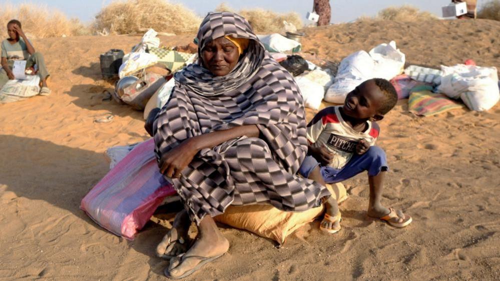 A woman wrapped in a cloth and wearing sandals looks down as she sits on a bag of her belongings, which is on sand. A small child is next to her and looks at her smiling.
