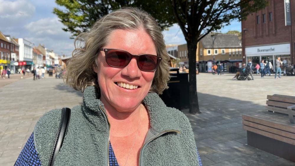 Sharon Dolan, a woman who is standing outside in Great Yarmouth and is wearing sunglasses over her eyes. She is smiling and looking directly at the camera and is wearing a blue blouse with a grey gilet over the top. 