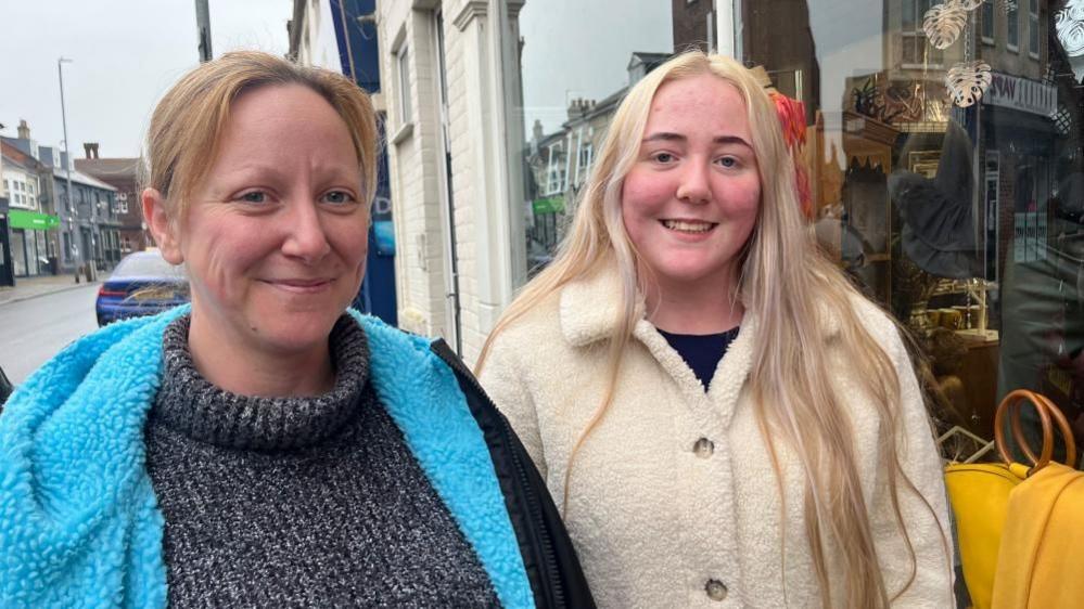 Bryony Marshall is on the left, in a blue-lined DryRobe coat and a grey flecked jersey. She has auburn hair. Her daughter, on the right, has long blonde hair, is smiling and is wearing a black top under a white fluffy coat. They are standing on Gorleston High Street. A yellow bag is visible inside the window of a café near where they are standing