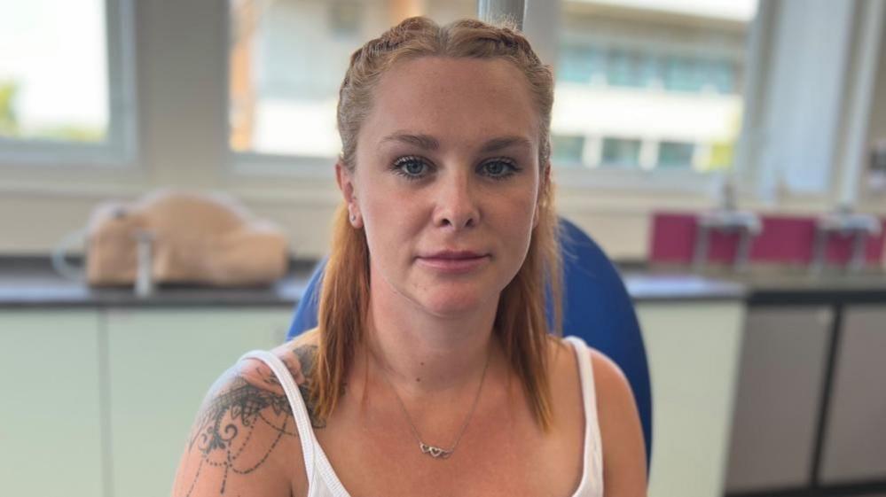 A woman with tied-back red hair looks straight at the camera. She is wearing a white strappy top and has a tattoo on her right shoulder.
