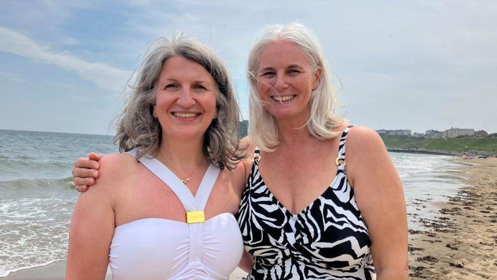 Two grey-haired women in swimsuits smile at the camera, a view of the beach behind them. The woman on the left has shoulder length wavy hair, and wears a white swimming costume with gold jewellery. The woman on the right has shoulder-length straight hair and wears a black and white patterned costume.
