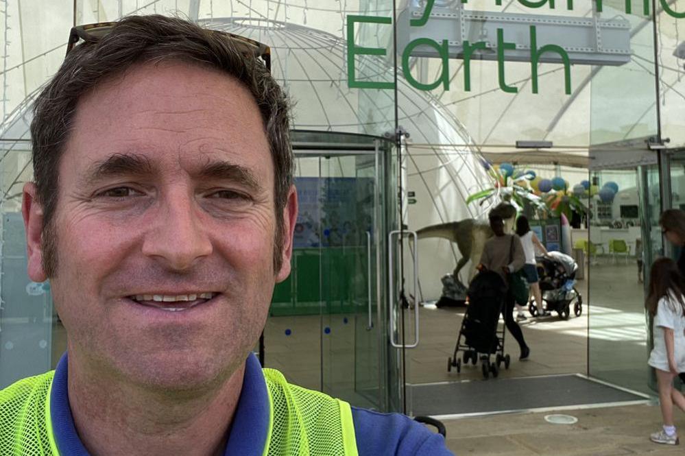 Ian takes a selfie in front of a building with floor to ceiling glass windows. He smiles and had short brown hair and wears a yellow and blue top.