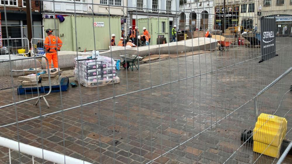 Fenced off paved area of market square with workers visible and a long concrete track which will form part of the water feature. It has clearly been raining.