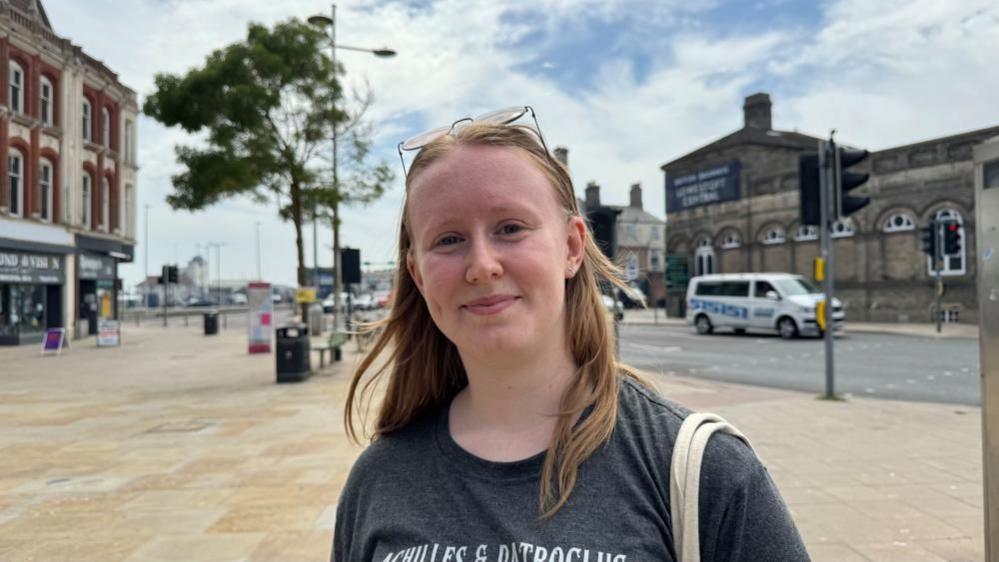 Katie Plant has long blonde hair, and her sunglasses are rested on her head. She is wearing a black tee shirt, and has bag straps over her left shoulder. She is pictured on the Station Square in Lowestoft, with Lowestoft Central railway station in the backdrop with the former Tuttles Department Store on the left of the image.