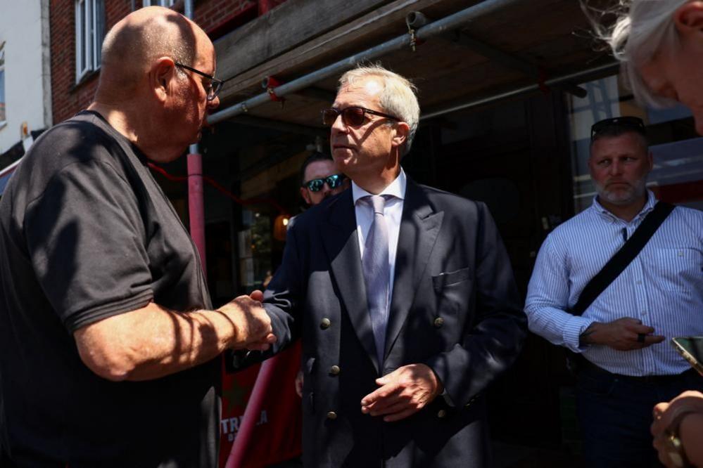 Farage in a navy blue suit shakes hands with a member of the public as another man behind him looks intently at the pair.