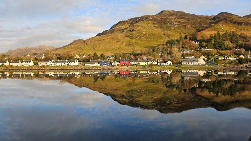 A bright, colourful and small rural village is reflected on the water. A hill sits behind the small town and it's a bright sunny day with some clouds.