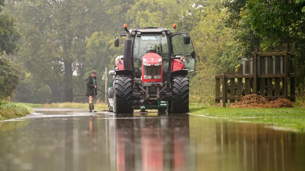 A man with a brown raincoat and wellington boots is standing in floodwater holding a rope attached to a car, which is not visible but is behind a red tractor. There are trees in the background.