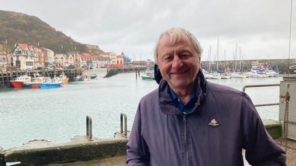 A man with white hair and a blue jacket and polo shirt standing in front of a bay with small boats and buildings in the distance.