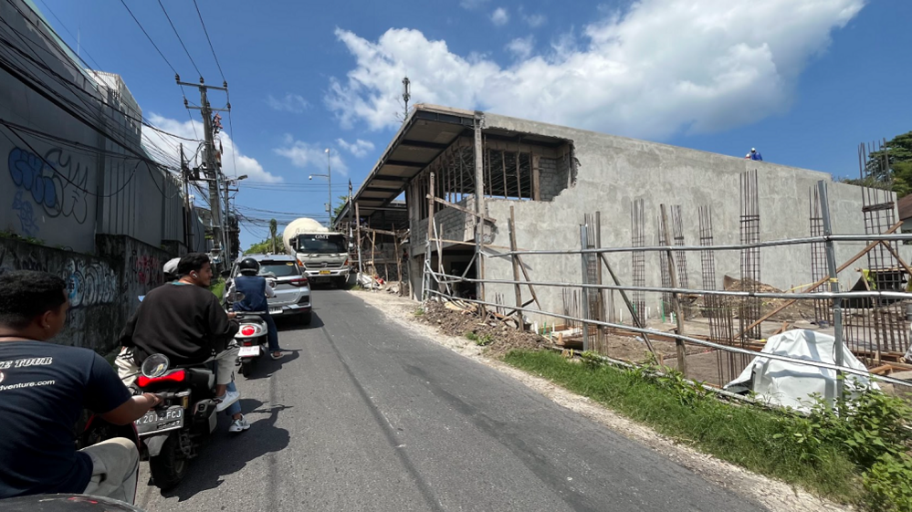 A line of motorcycles waiting along a narrow road as a car is trying to squeeze past a large truck at a construction site.