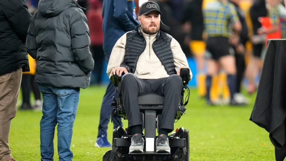 Former Gloucester Rugby player Ed Slater after the final whistle in The 745 Game at AMT Headingley Stadium, Leeds. He is wearing a black 4Ed hat, a black gilet, a beige fleece and black trousers. Ed is sat in a wheelchair on the pitch. Other people can be seen standing around him and nearby.