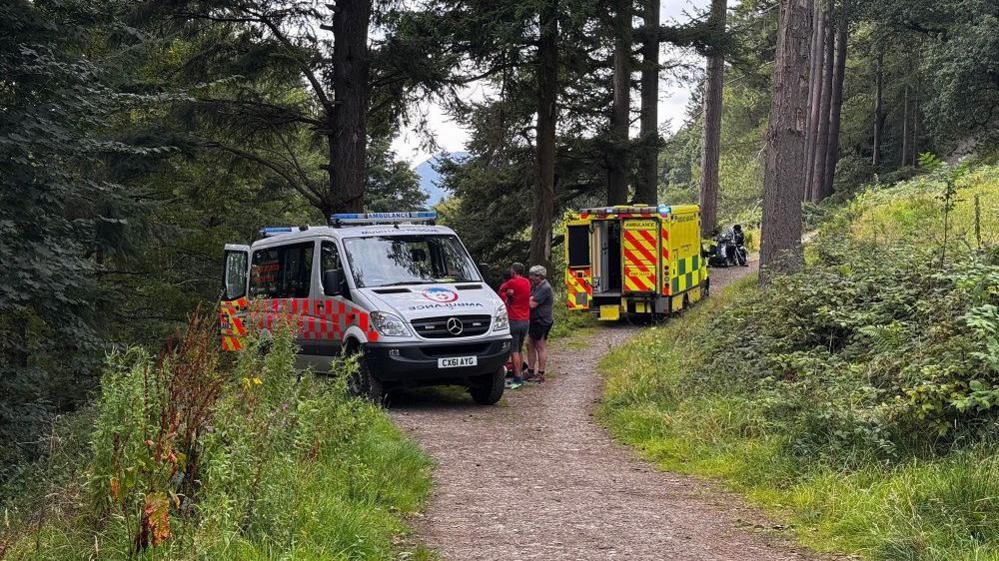 The mountain rescue van and ambulance parked on a path surrounded by trees. The back doors to the van are open and people are gathered to the side of it. The back door to the ambulance is also open.