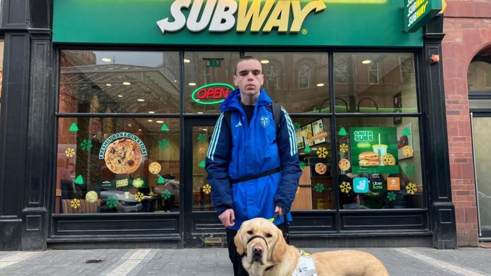 A teenager with short dark hair and a neutral expression stands with a golden Labrador guide dog in front of a Subway shop. He wears a blue Leeds United coat and holds a lead connected to a high-visibility harness on the dog. The green and yellow 'Subway' sign is illuminated above his head and the window display advertises cookies and sandwiches.