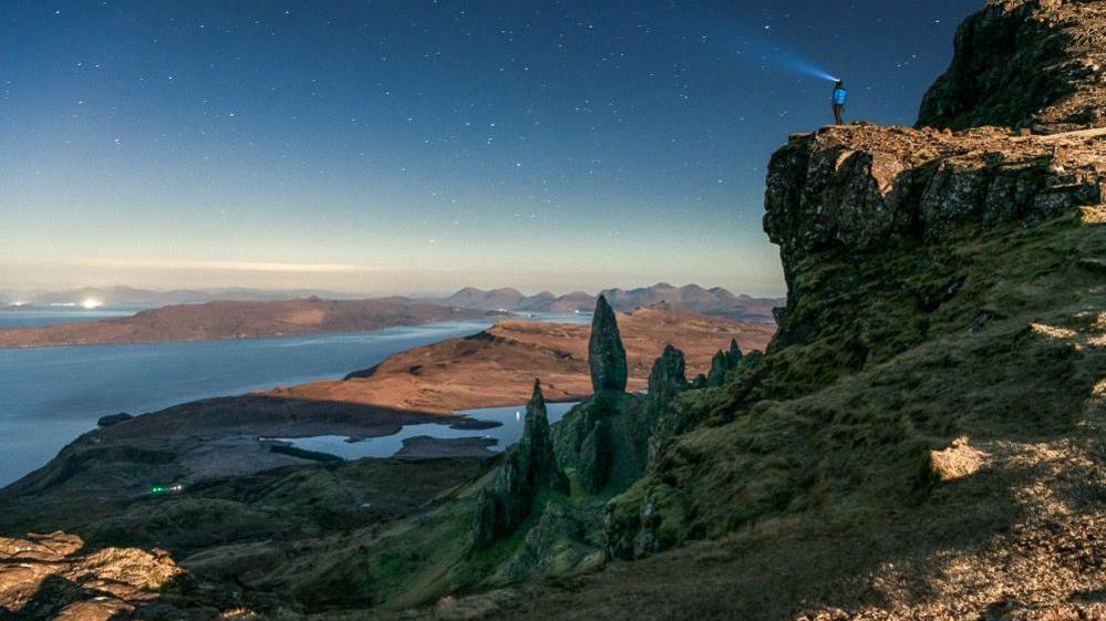 Landscape image of a person standing on the edge of a cliffside with trees and a loch below a steep incline. The sky is a gradual blue gradient with stars visible.