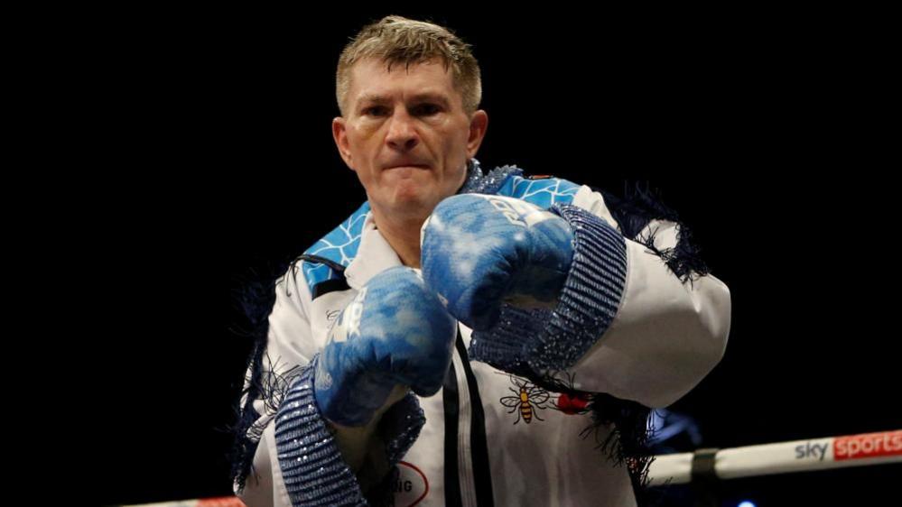 Ricky Hatton stands in a boxing ring. He is wearing a white tasselled suit and blue boxing gloves and punching the air. 