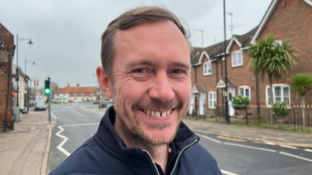 Adam Blake, with mousy brown hair and goatee beard, is smiling by the roadside through Acle. He is wearing a blue jacket.