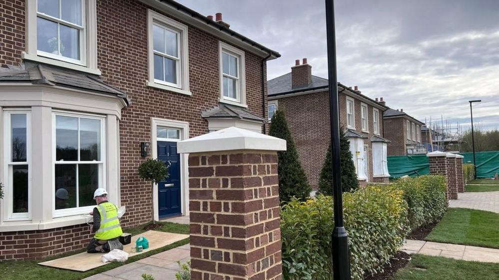 A photograph of the first show home showing hedges at the front and a man in a hard hat working on the windowsill. The house has sash windows, a blue front door and red brick walls.