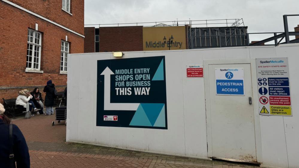 White hoardings with a large sign that says "Middle entry, shops open for business this way" with an arrow next to it. To the left of the shot a red brick building can be seen, with a group of women sitting on a bench in front of it