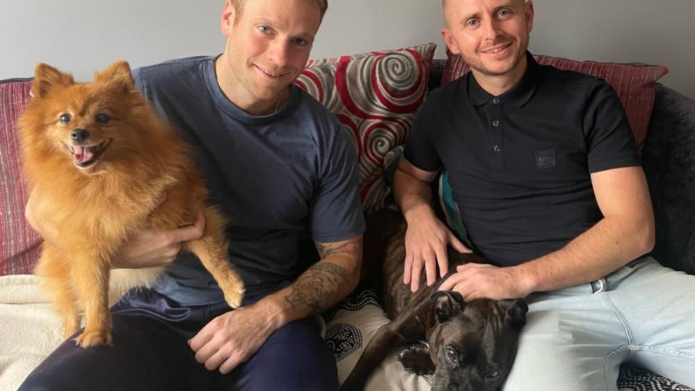 Two men sit on a couch. The one of the left is holding a brown Pomeranian dog and the one on the right is patting a black boxer dog. 