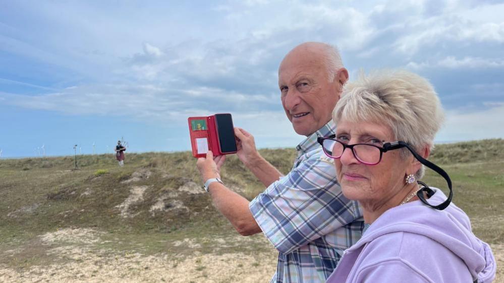 David Angel is wearing a blue checked shirt, with short sleeves. He is holding his mobile phone, in a red case with green bank card showing. He has a wrist watch on his left arm, and has close cropped white hair, balding on top. Pat has medium length blonde hair, wears pink-rimmed glasses and is wearing a mauve hoodie. In the distance is piper Ryan Fitzpatrick playing his bagpipes.