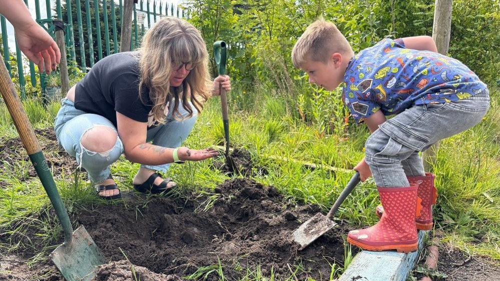 Sam, who has brown hair is showing her son something small she found in the clay in her hand. Chester is wearing red wellies and digging with a shovel in the park.