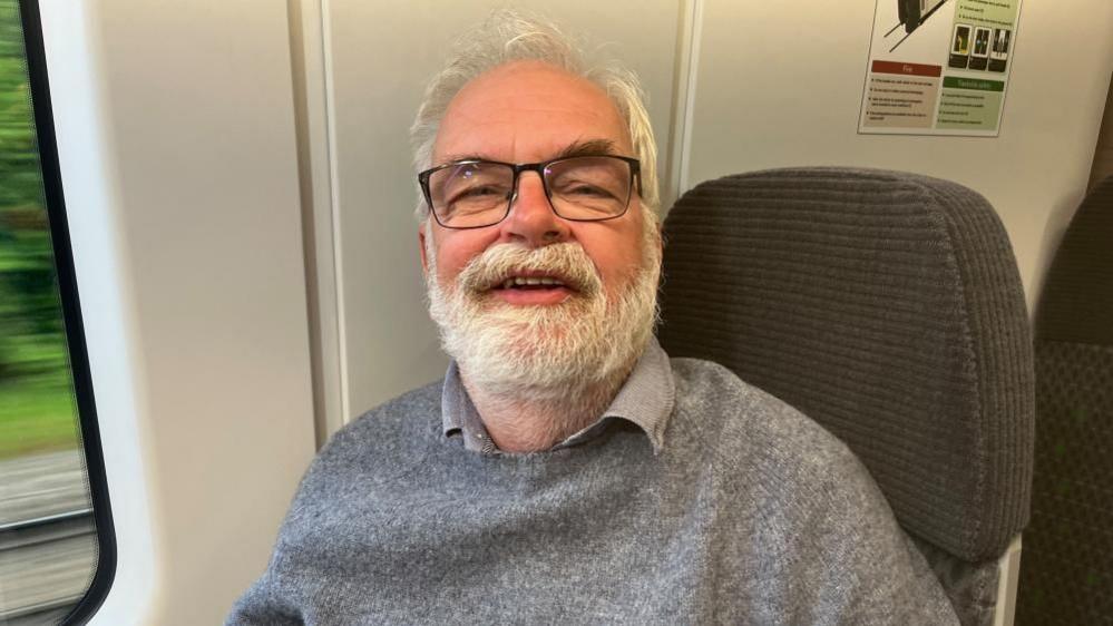 Phil Warner, sitting in a railway carriage, wearing a grey jumper and shirt. He is looking at the camera, smiling, he has a grey beard, dark glasses and short grey hair.